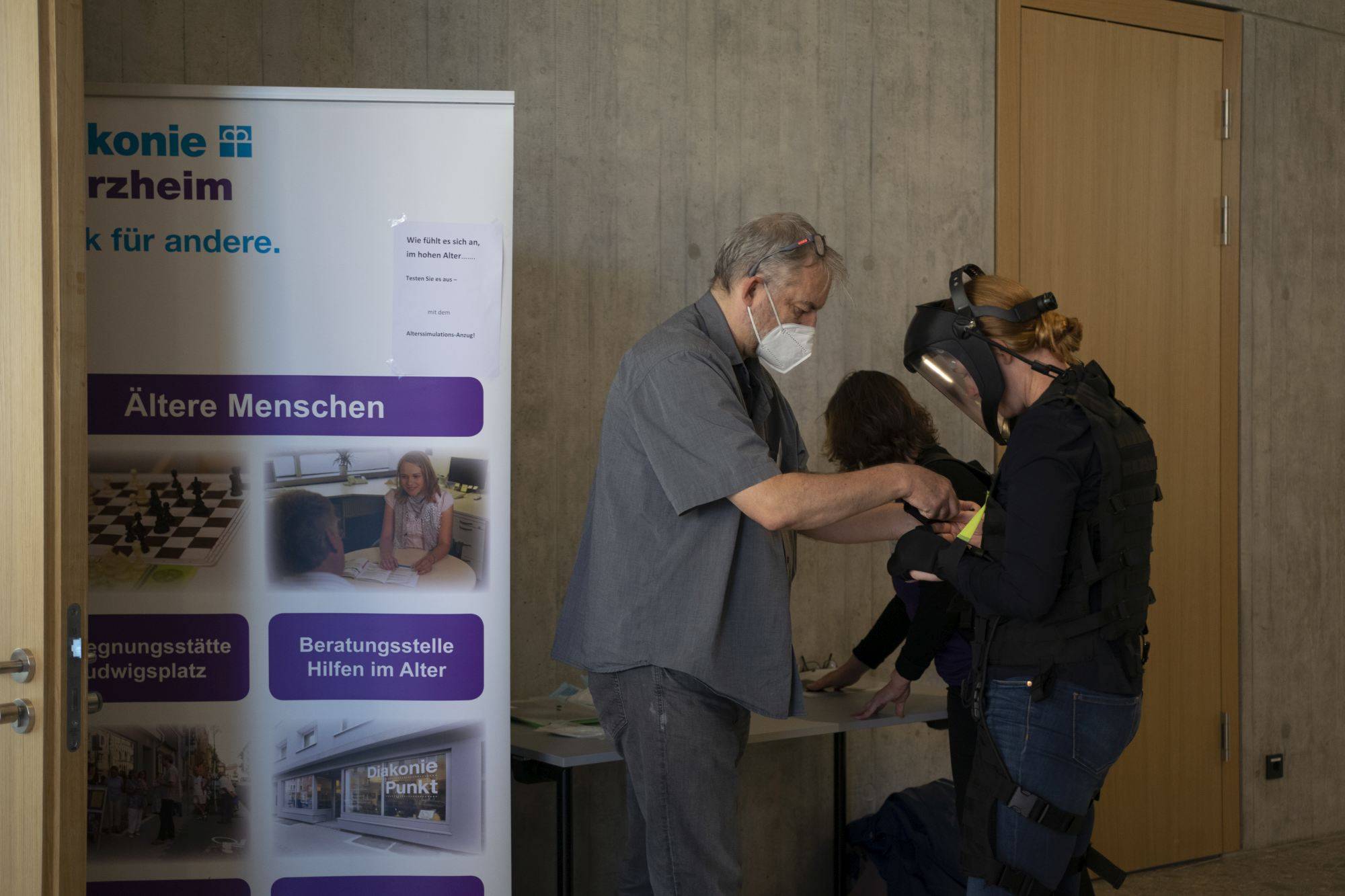 Quelle: ©EvKirchePF ein Mann zieht einer Frau einen "Alterssimulationsanzug" an